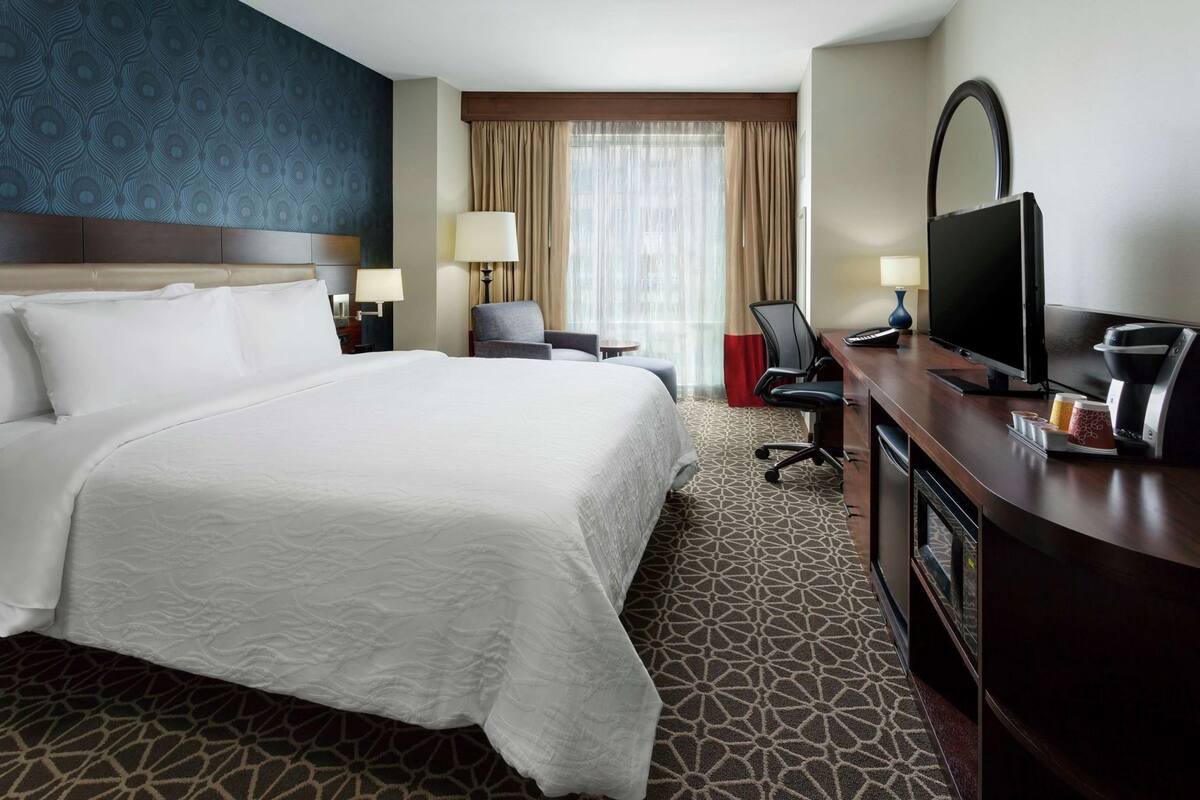 A hotel room with a large bed, a desk with a TV, a chair, a lamp, and a window with curtains.