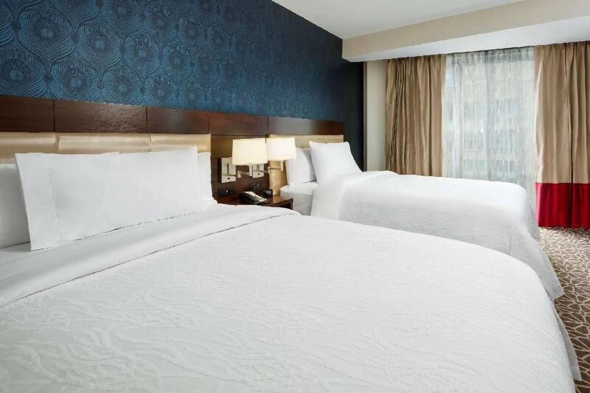 A hotel room with two beds, a headboard, a nightstand with lamps, and a window with curtains.