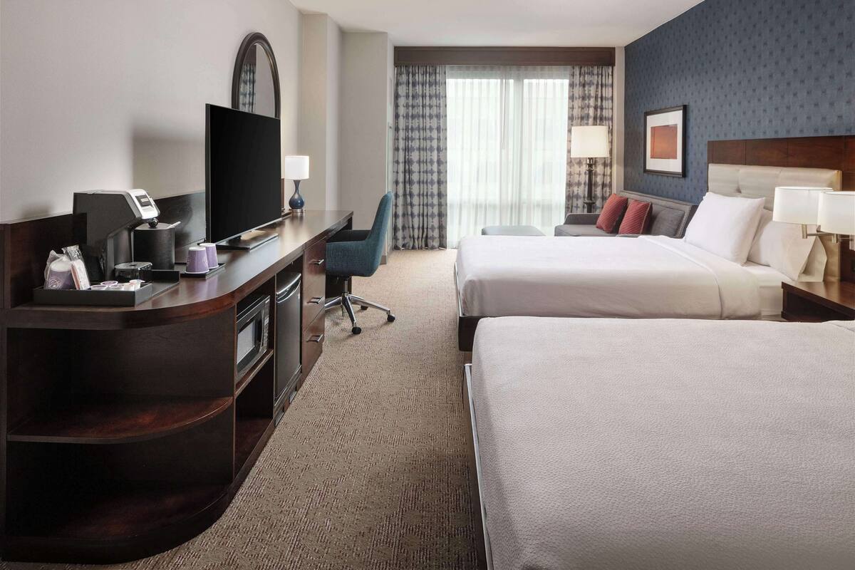 A hotel room with a desk, television, two beds, and a large window with curtains.