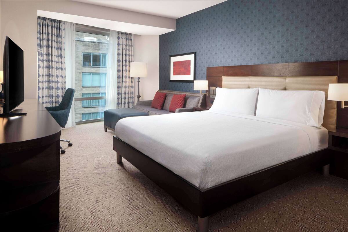 A hotel room with a large bed, a desk with a TV, a chair, and a view of the city through large windows.
