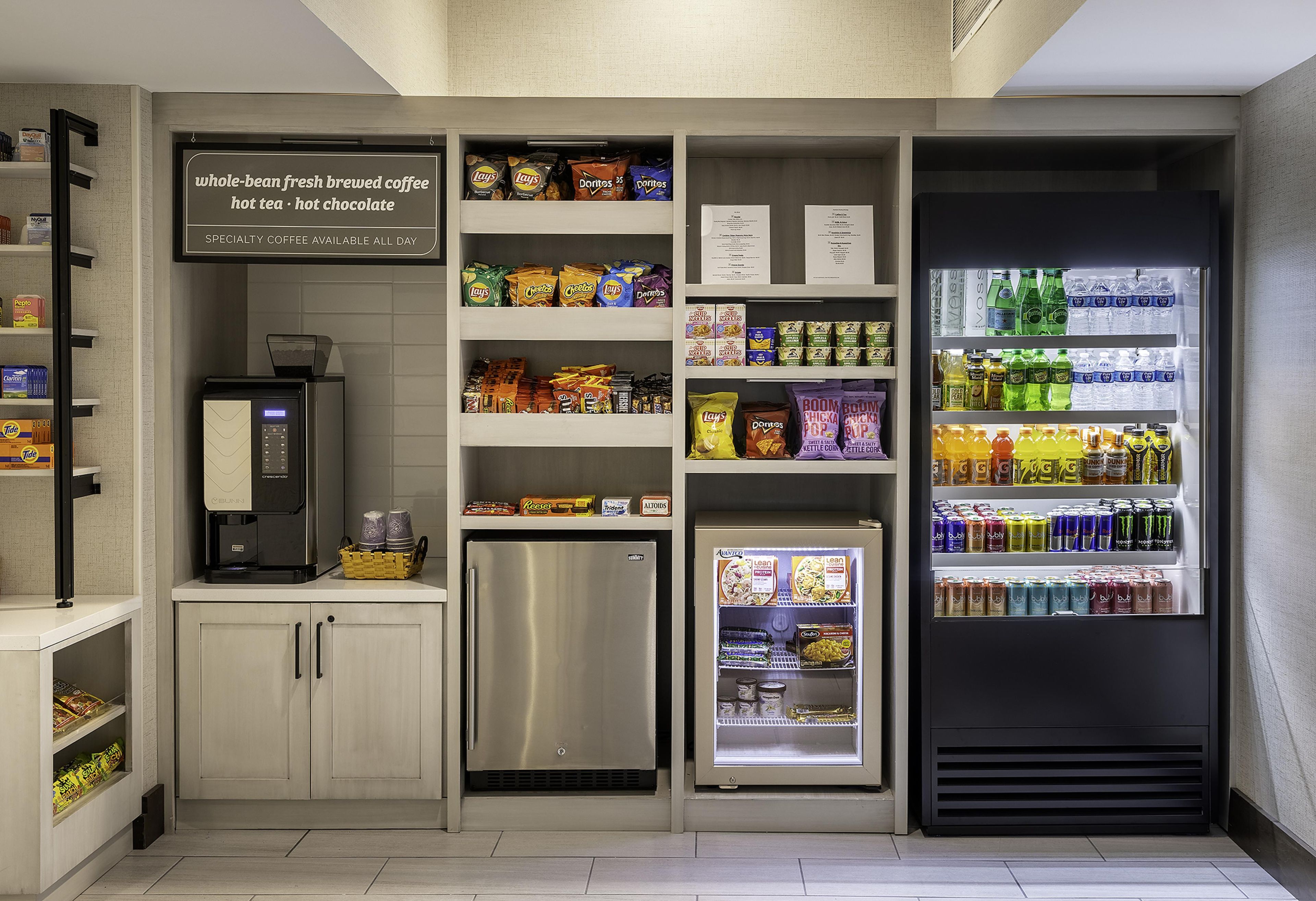 A kitchenette with a coffee machine, refrigerator, and shelves stocked with snacks and beverages.
