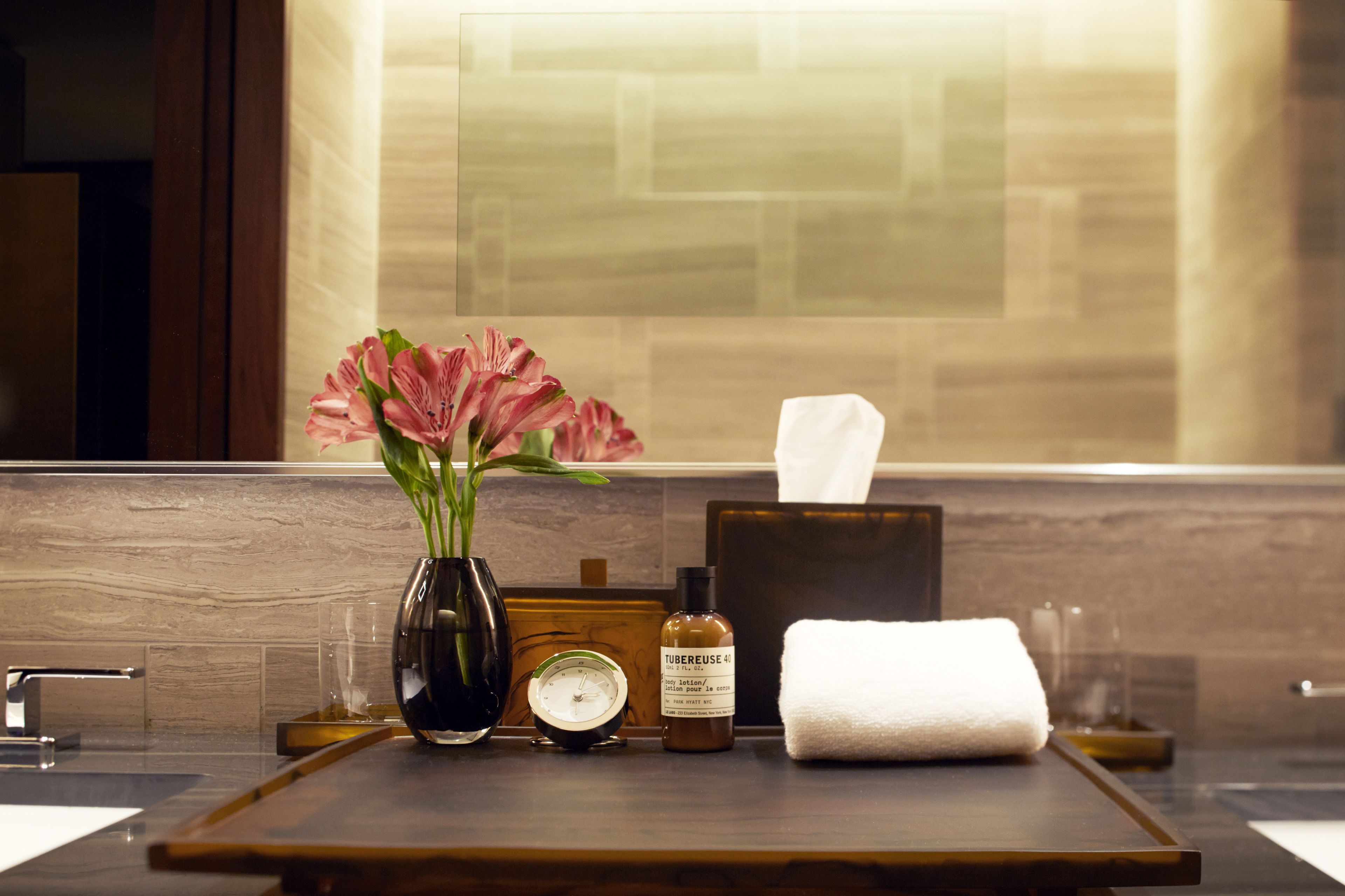 A bathroom vanity with a vase of pink flowers, a tissue box, a bottle of lotion, and a clock.