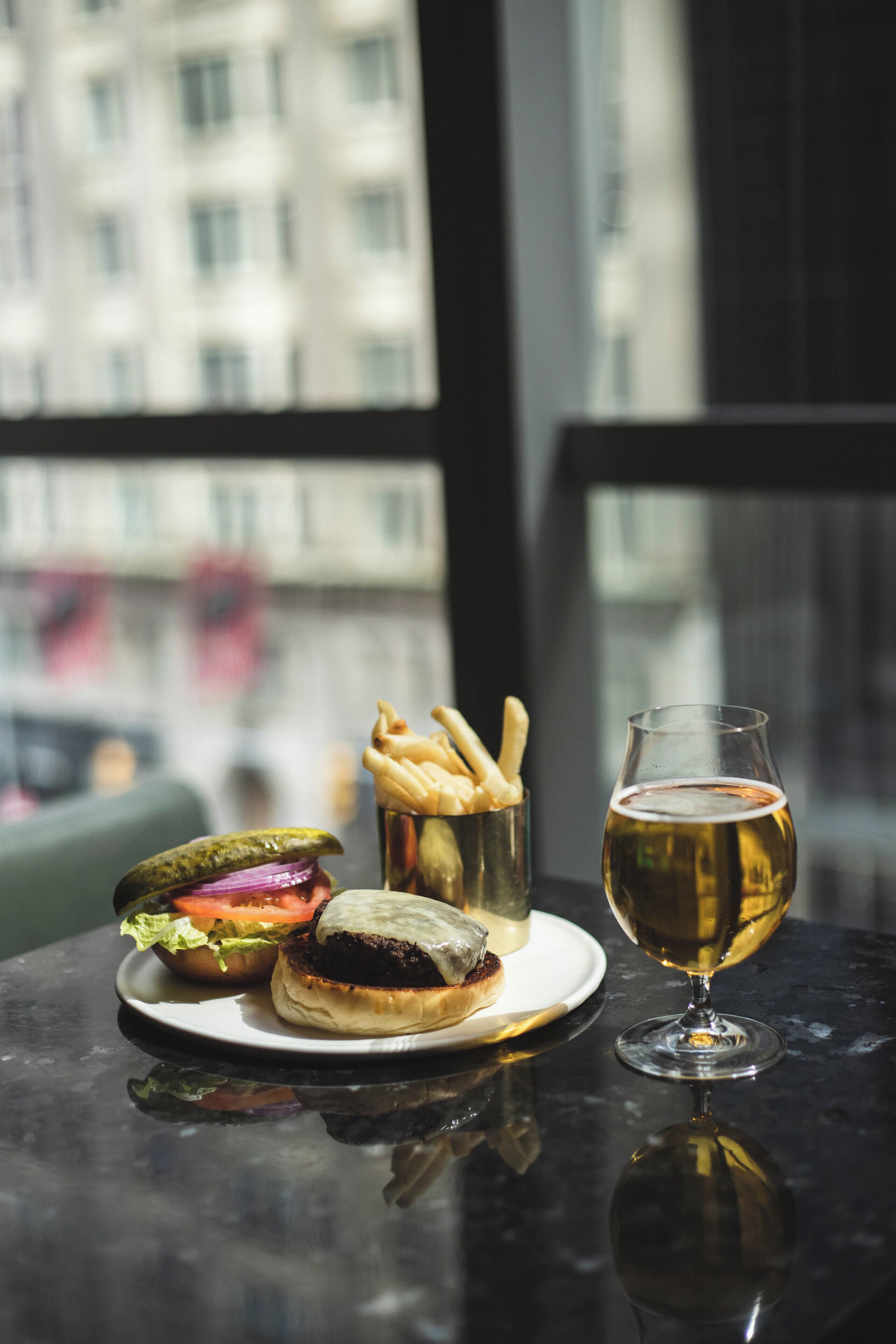 A table with a burger, fries, and a glass of beer, with a city view in the background.