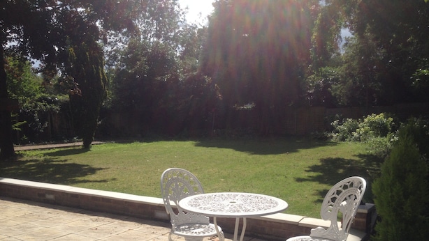 Une terrasse avec deux chaises en métal blanc et une table, entourée d’un jardin avec une pelouse et des arbres.