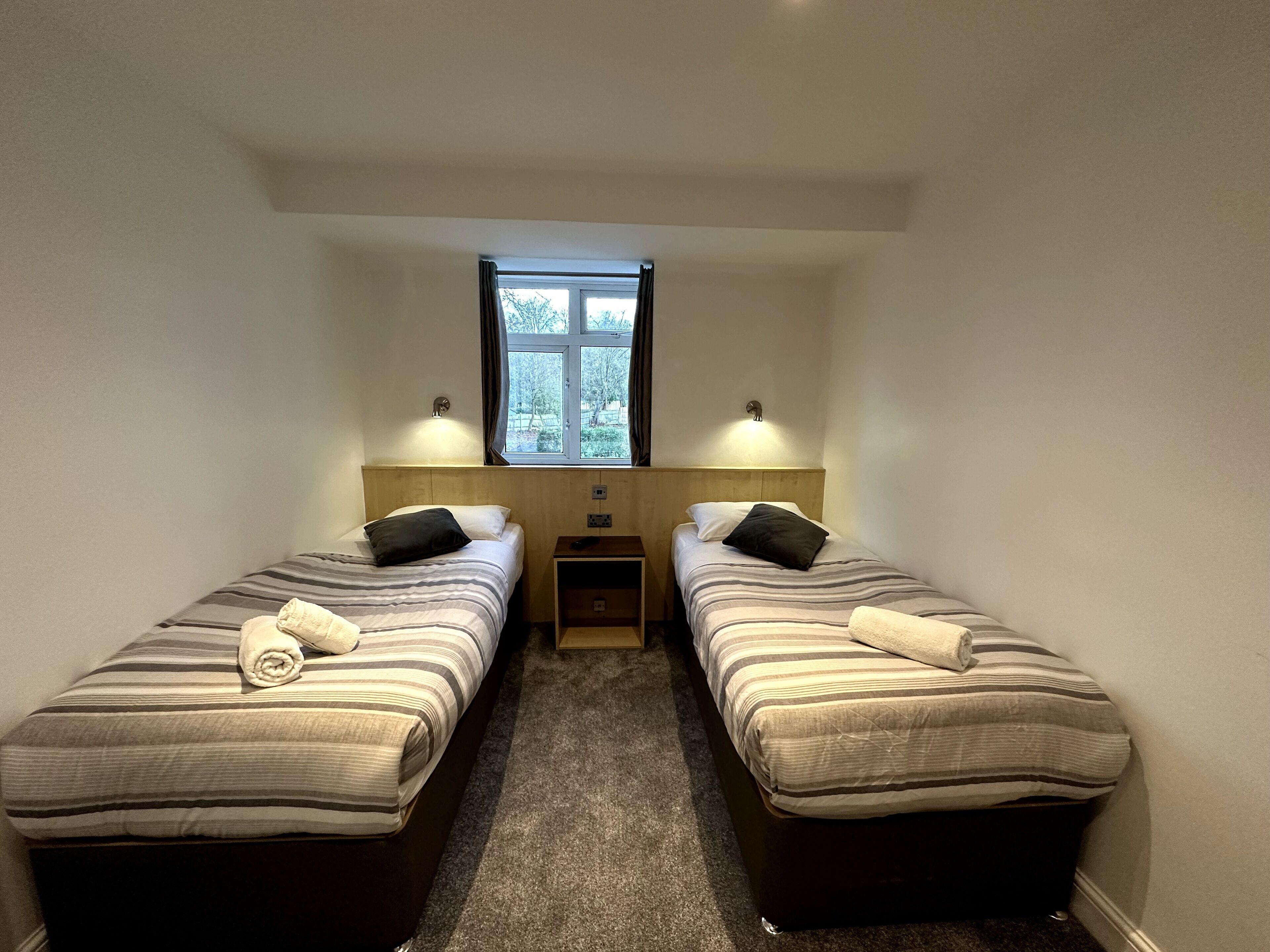 A small, well-lit room with two single beds, a window, and a nightstand between them.