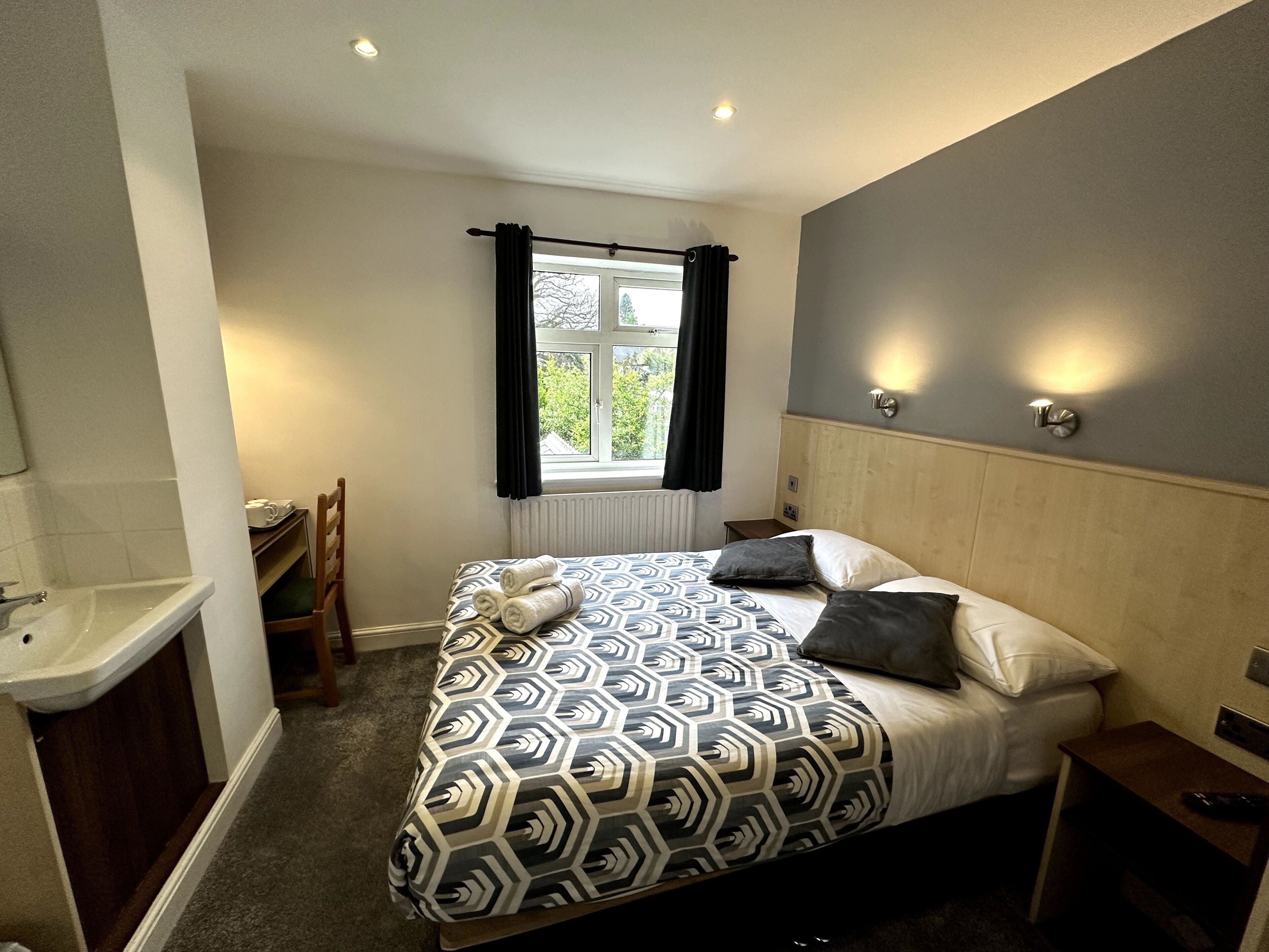 A hotel room with a bed, a bedside table, a chair, a window with curtains, and a bathroom with a sink.