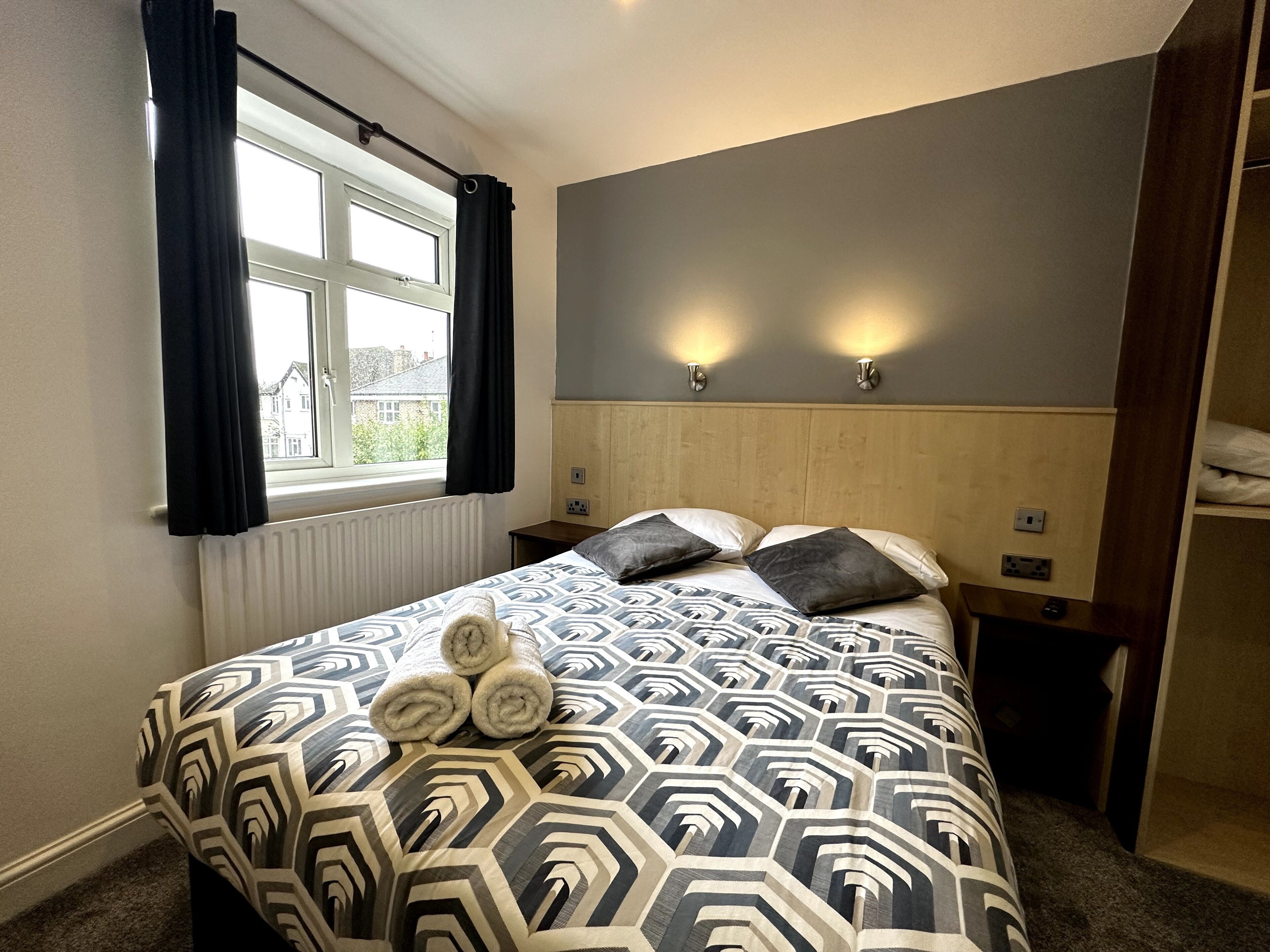Quarto de hotel com cama, janela com cortinas, radiador e duas luzes fixadas na parede.