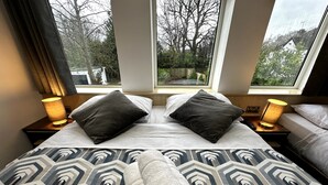 Une chambre d’hôtel avec un grand lit, deux lampes de chevet et une vue sur des arbres et une maison, visible par les fenêtres.