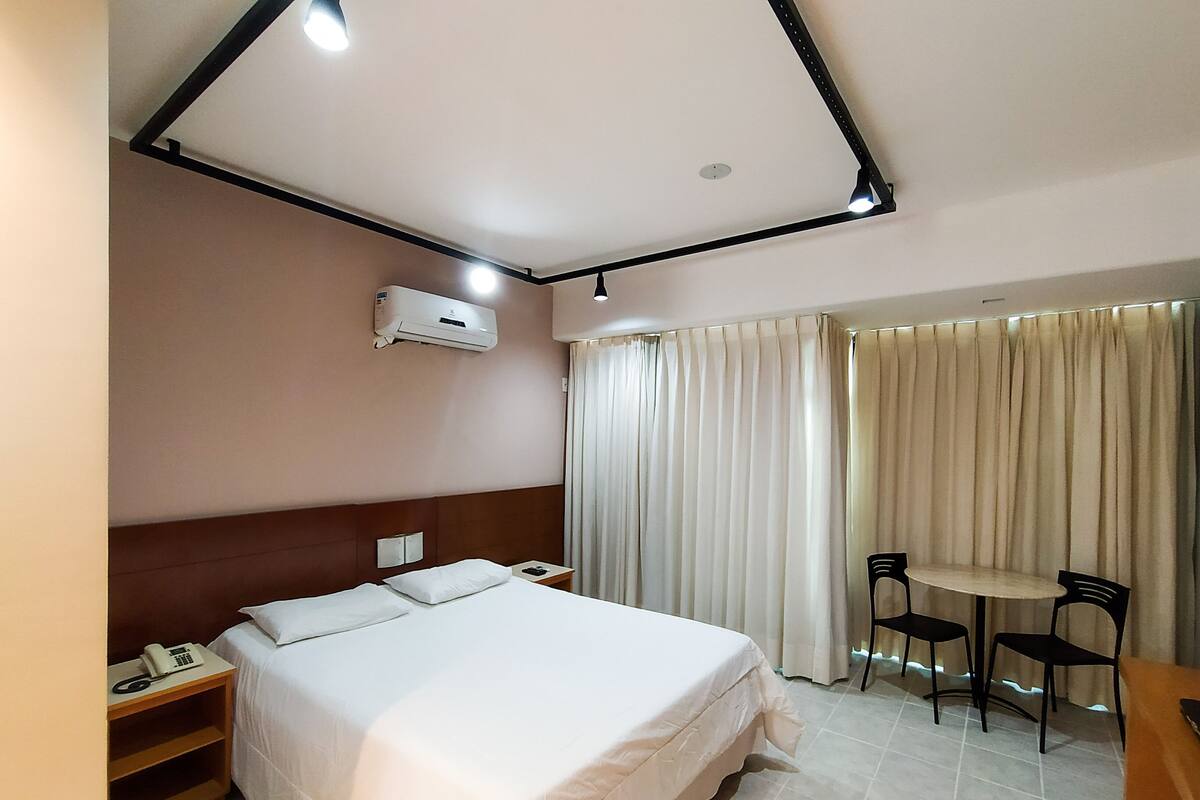 Habitación de hotel con cama, mesita de noche, teléfono, equipo de aire acondicionado, cortinas y una pequeña mesa con sillas.