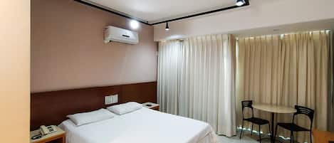 Quarto de hotel com cama, mesa de cabeceira, telefone, ar condicionado, cortinas e uma pequena mesa com cadeiras.