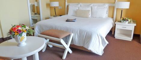 A hotel room with a bed, a bedside table, a chair, a small table with a vase of flowers, and a mirror.