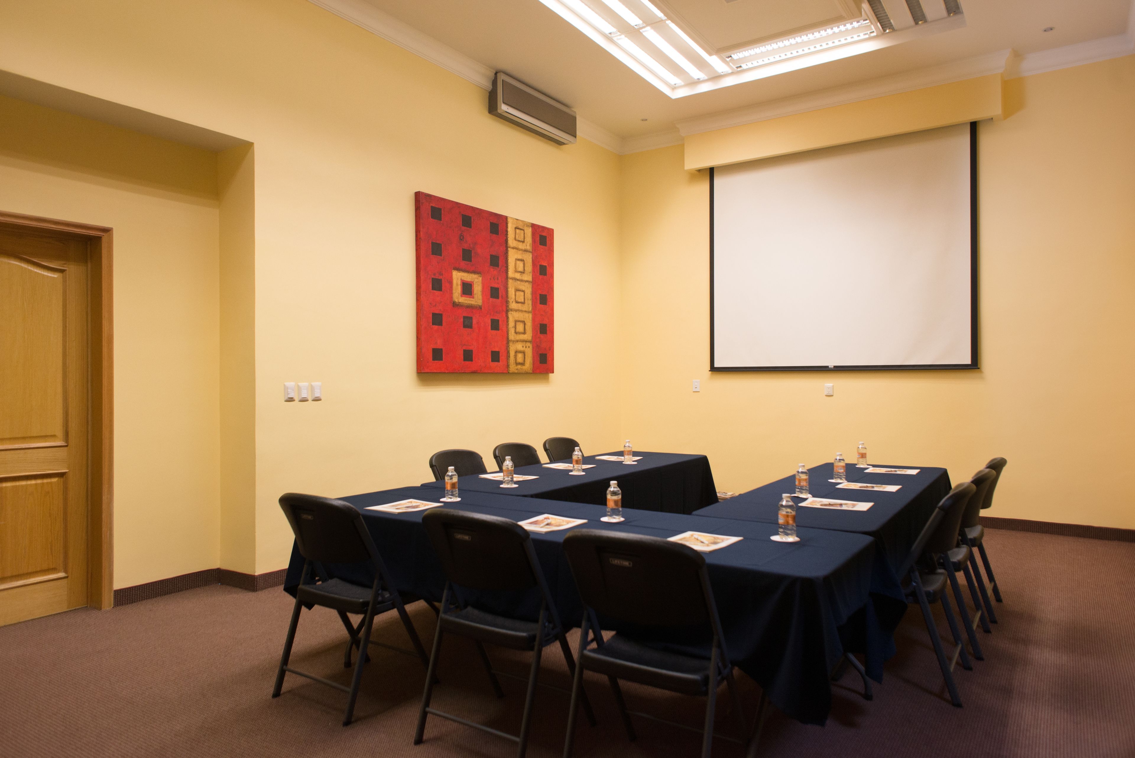 Un salón de conferencias con una pantalla de proyector, un cuadro abstracto rojo y una puerta de madera.
