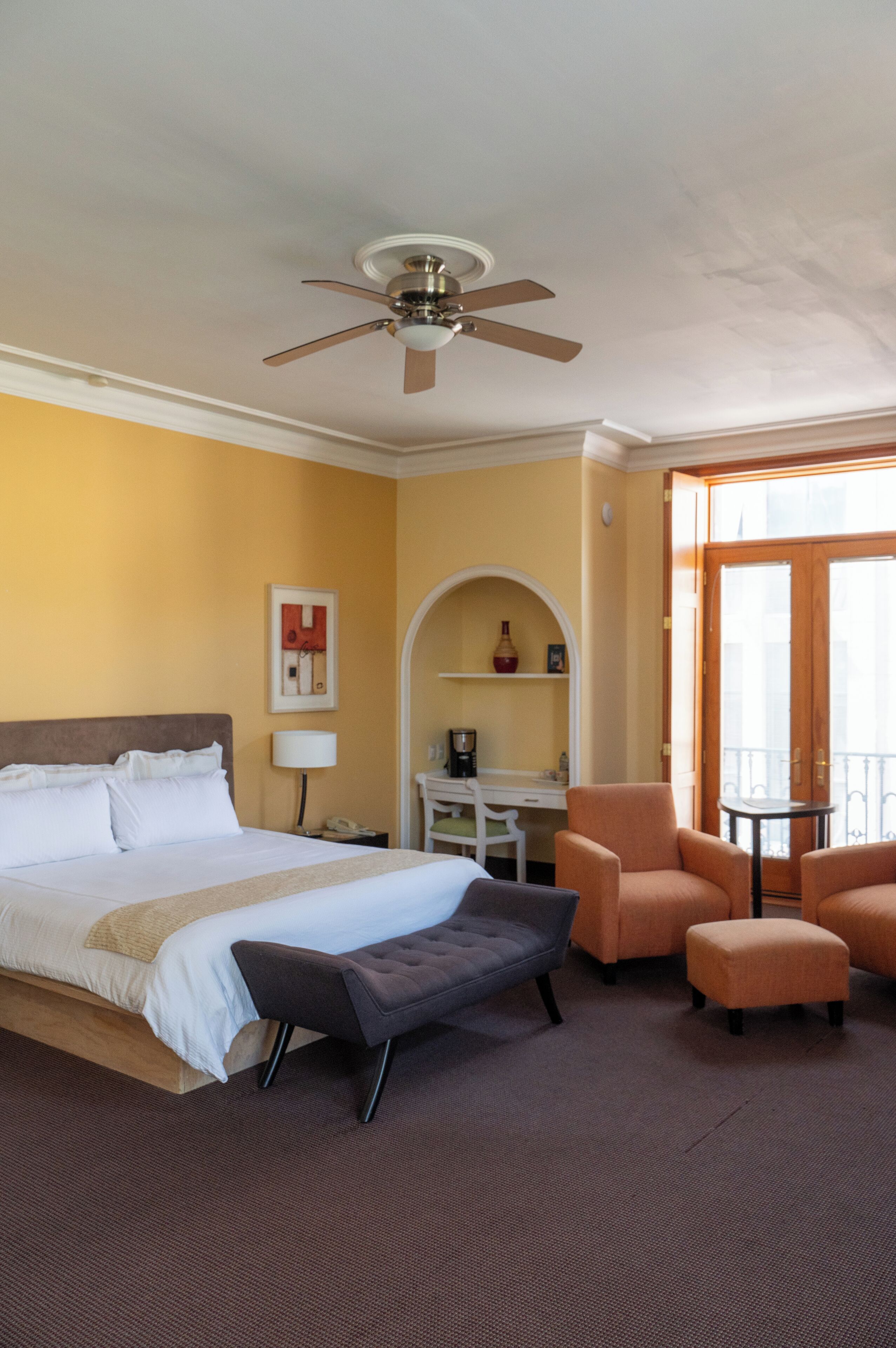 A hotel room with a bed, a desk, a chair, an ottoman, a ceiling fan, and a window.