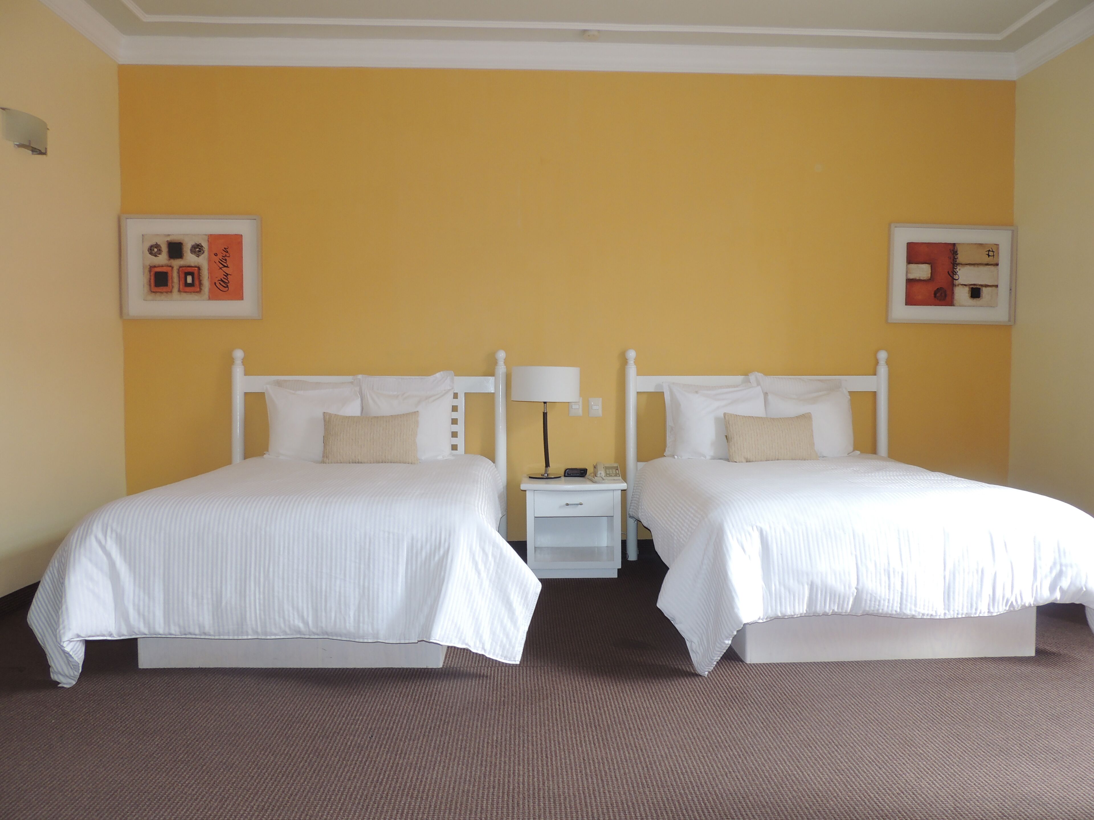 A hotel room with two single beds, a nightstand with a lamp, and two framed pictures on the walls.
