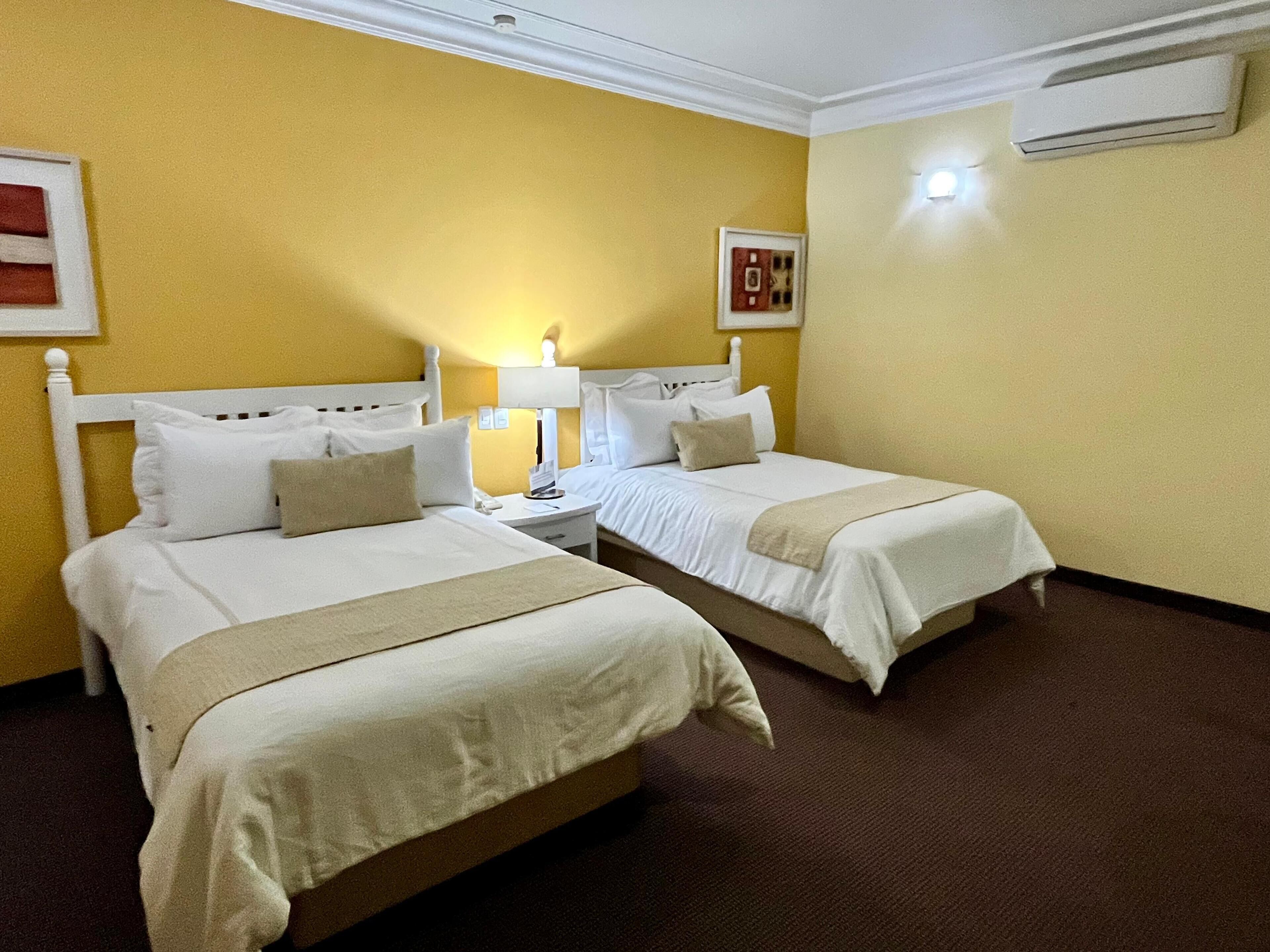 Habitació d'hotel amb dos llits individuals, una tauleta de nit amb una làmpada, aire condicionat fixat a la paret i un quadre encimbellat.