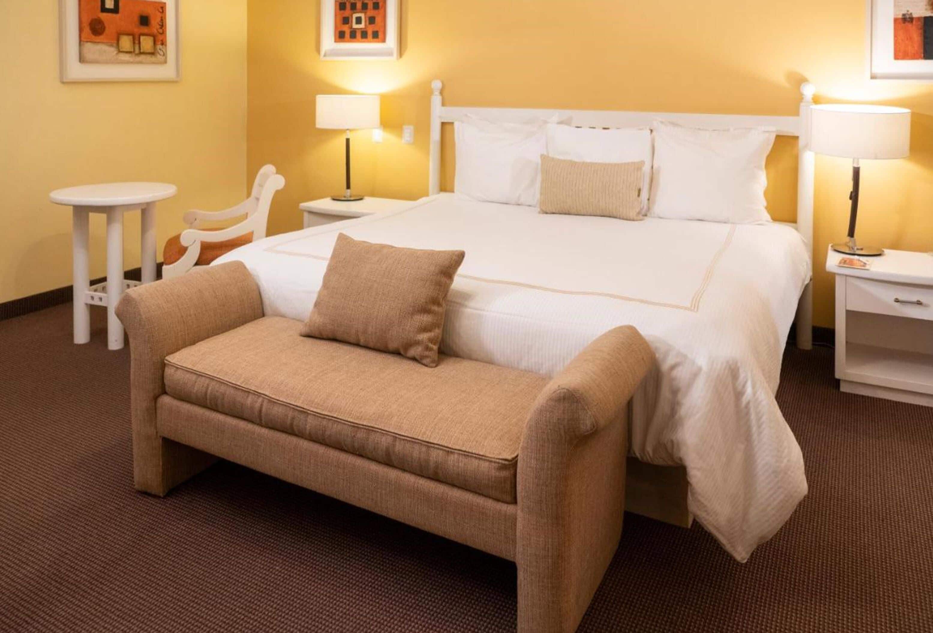 Quarto de hotel com uma cama grande, um sofá pequeno, uma mesinha de cabeceira, uma luminária e uma cadeira.