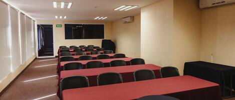 A conference room with rows of tables and chairs, a projector screen, and an air conditioning unit.