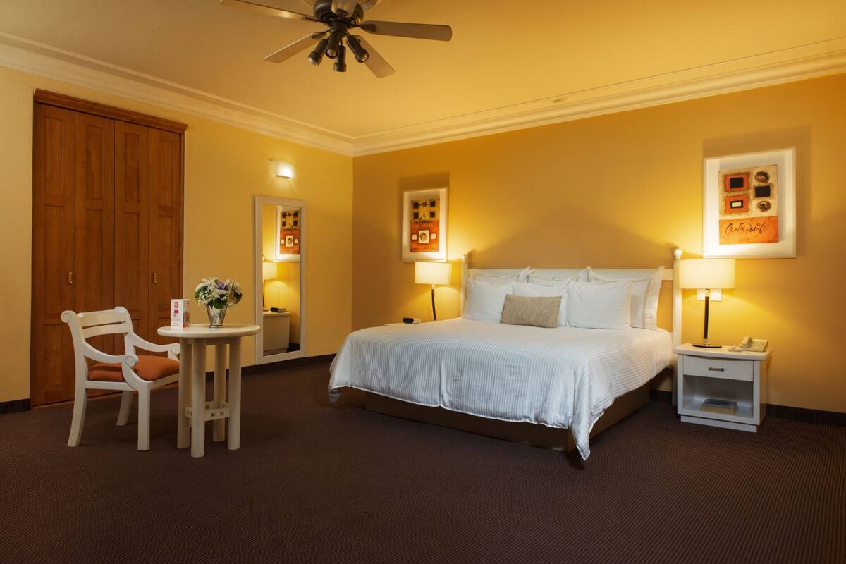 A hotel room with a bed, bedside tables, a chair, a ceiling fan, and wall-mounted artwork.