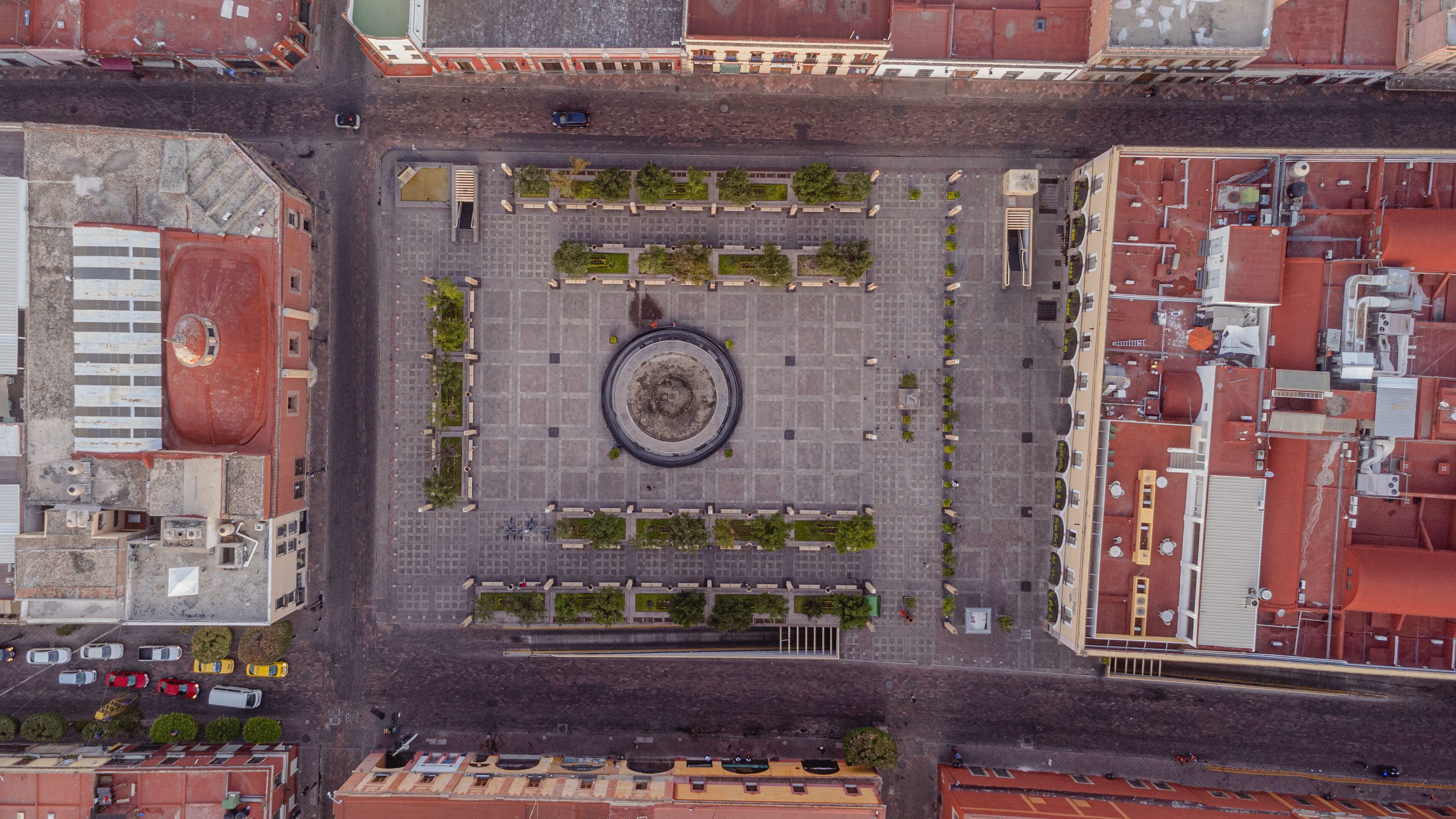 Vista aérea de una plaza rectangular con un monumento circular central, rodeado de árboles y bancos distribuidos uniformemente.