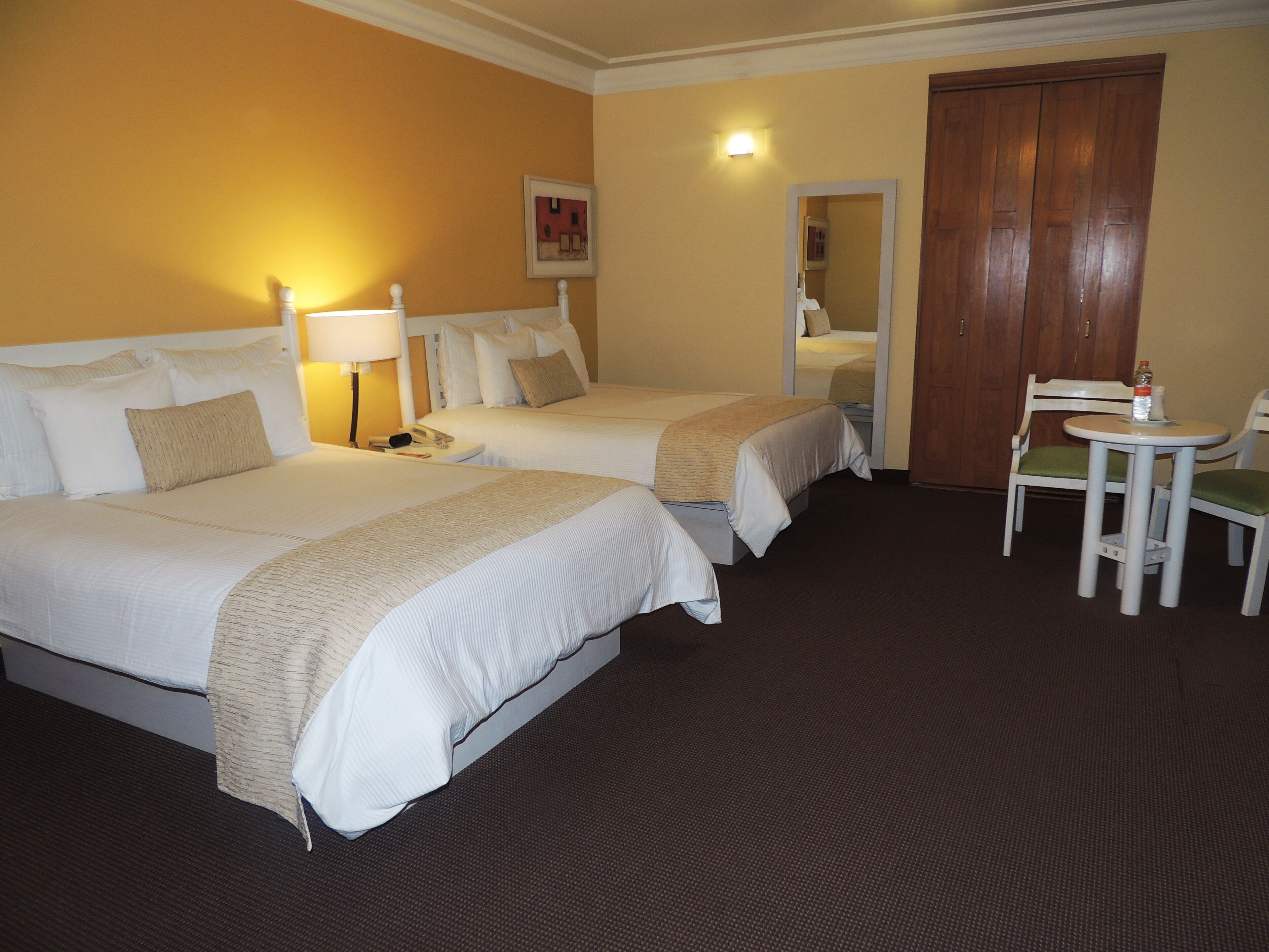 A hotel room with two beds, a small table with chairs, a lamp, and a wooden door.