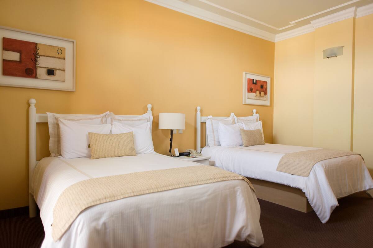 A hotel room with two single beds, a bedside table with a lamp, and artwork on the walls.