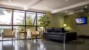 A hotel lobby with a large window, a sofa, two armchairs, a small table, and a television on the wall. - Guara Inn (Guaratingueta)