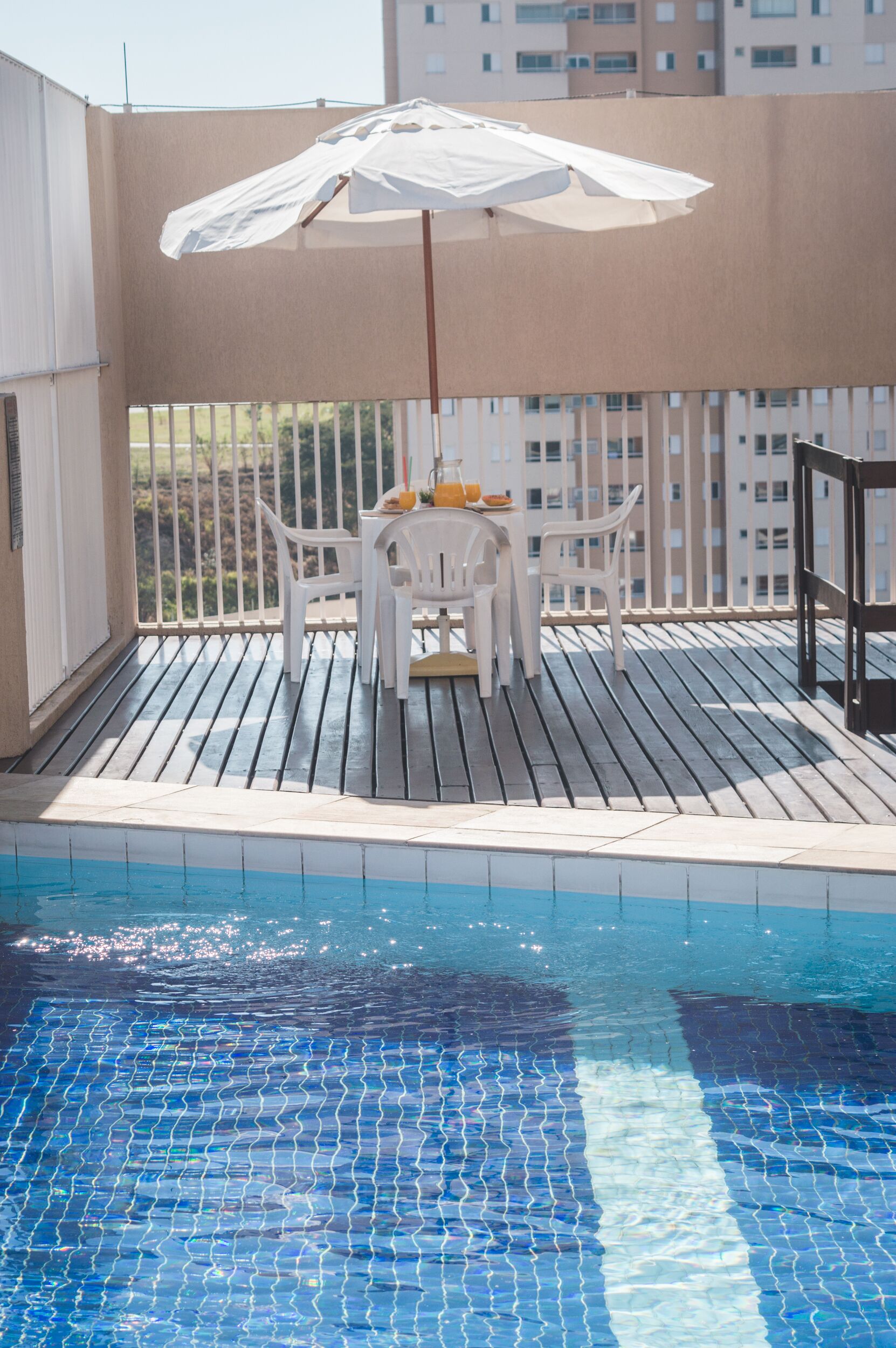 A poolside area with a table and chairs under a white umbrella, surrounded by a wooden deck and a pool with blue tiles.