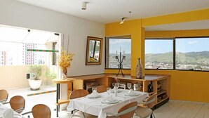 A dining area with a table set for four, a potted plant, and a view of the city through large windows. - Guara Inn (Guaratingueta)