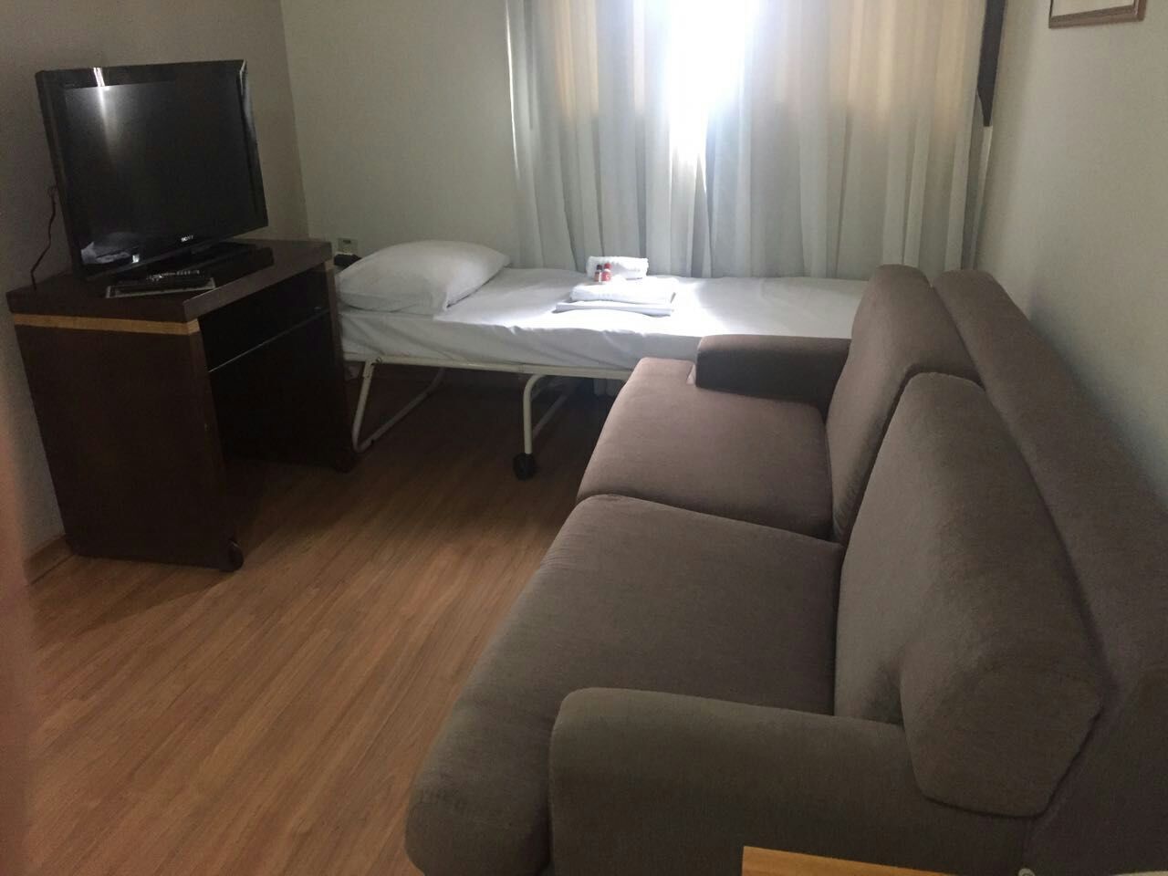 Een kleine kamer met een bank, een televisie op een standaard, een bed met een witte matras en kussens, en een raam met gordijnen.
