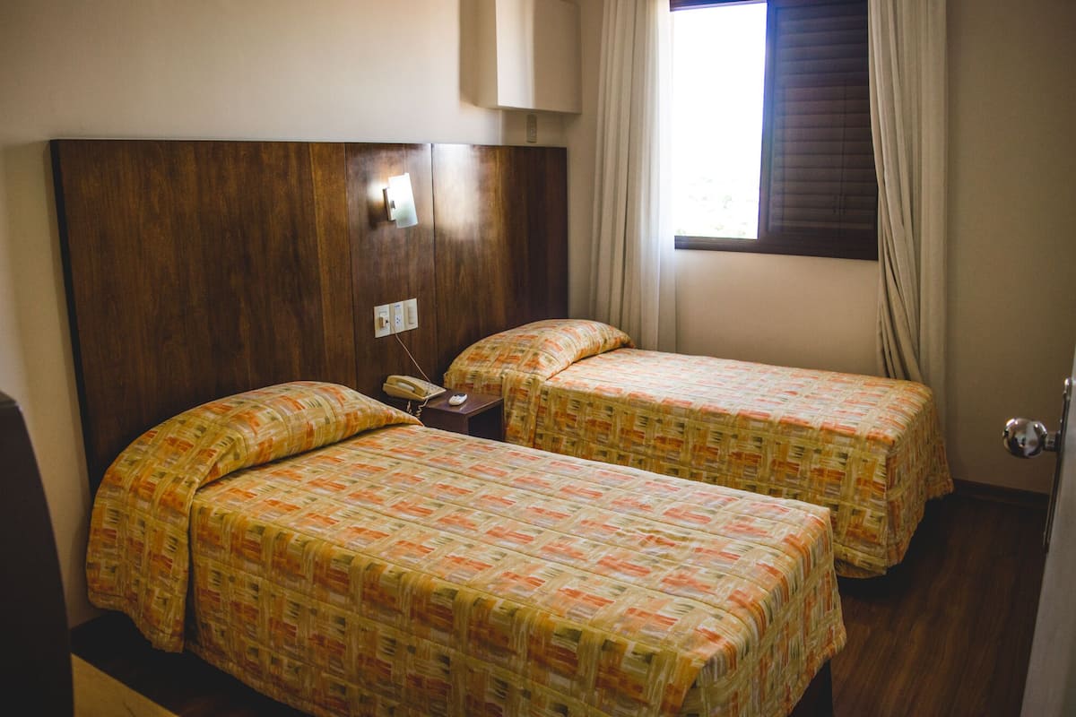 Quarto de hotel com duas camas, cabeceira de madeira, telefone ao lado da cama e janela com cortinas.