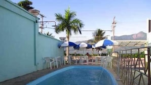 Una piscina con cerramiento metálico, sillas blancas y sombrillas.