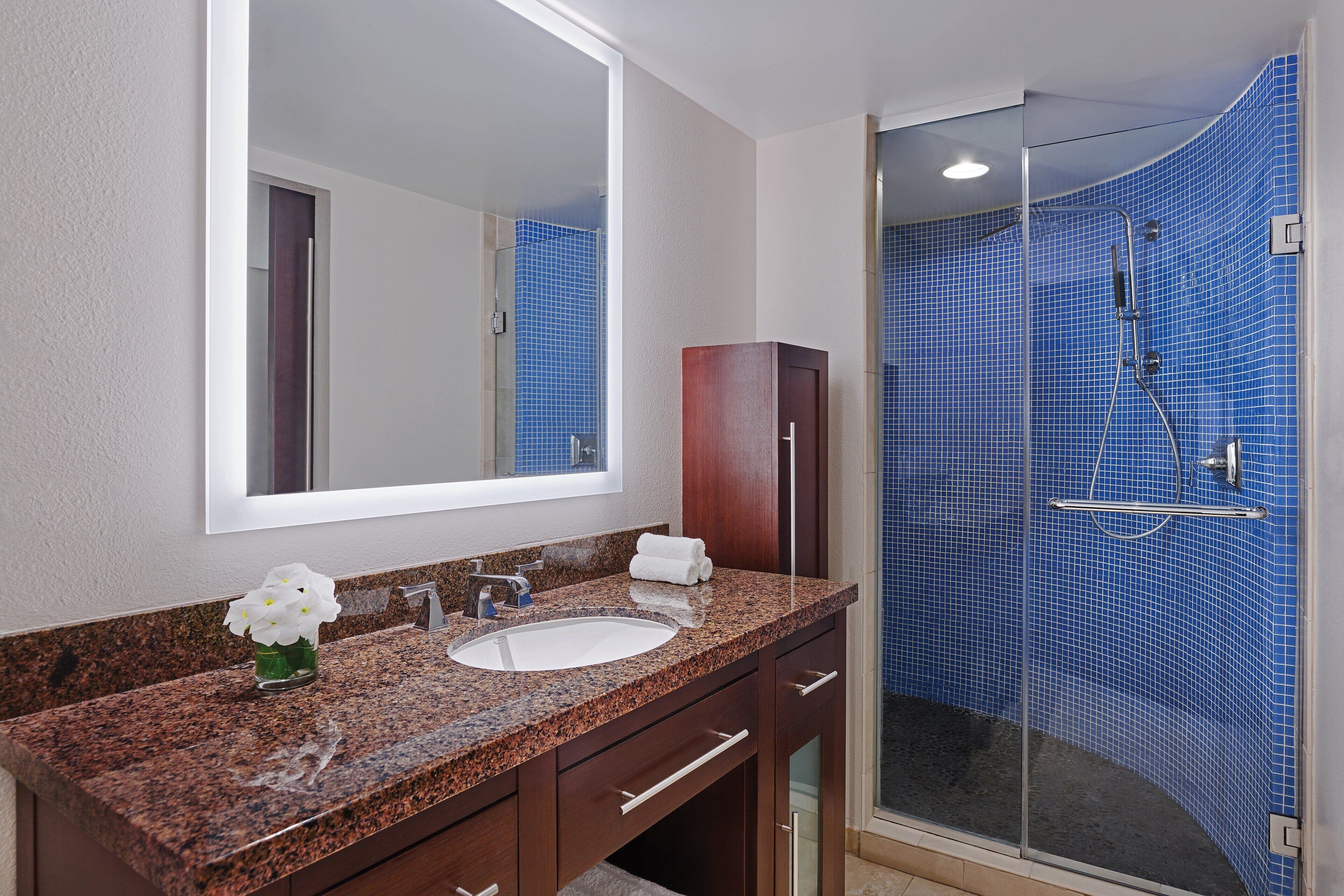 A bathroom with a granite countertop, a white sink, a mirror, a wooden cabinet, and a glass-enclosed shower with blue tiles.