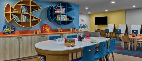 Zona de juegos infantiles con una mesa redonda, sillas azules y estanterías de madera llenas de juguetes y libros.