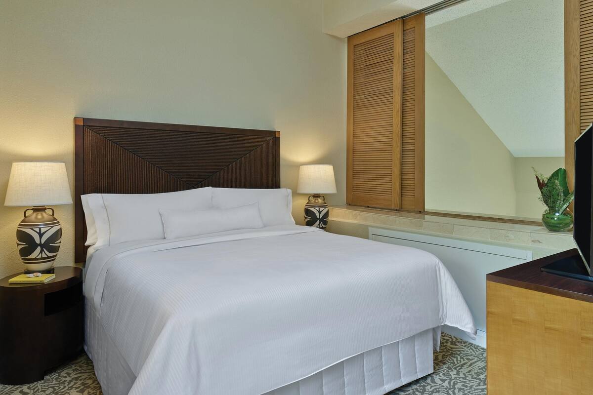 Una cama bien hecha con cabecera de madera, dos lámparas de noche y un televisor sobre un mueble de madera.