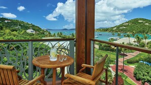 Una terraza de madera con mesa y sillas, con vista al mar y barcos, rodeada de vegetación.