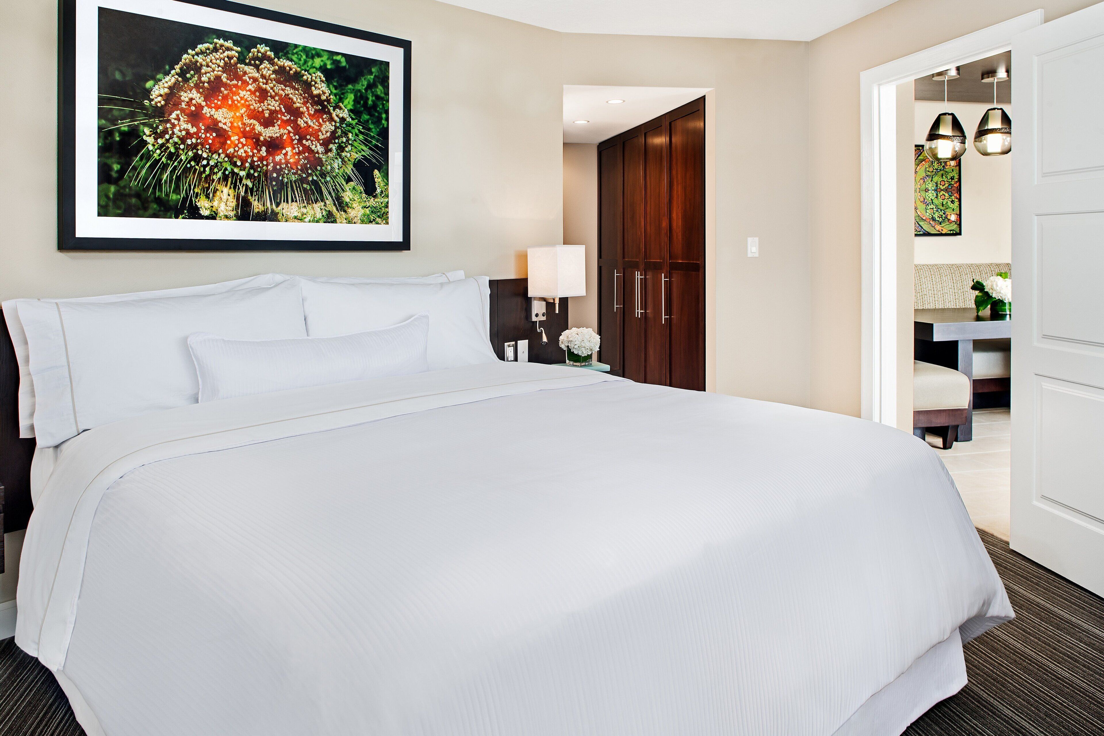 Een hotelkamer met een groot bed, een lijst met bloemenkunst boven het bed, een bureau met een lamp en een zichtbare badkamer door een open deur.