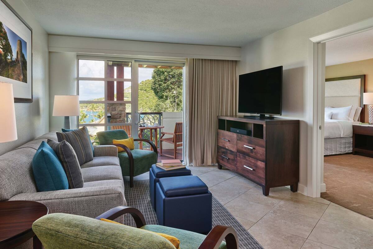 Habitación de hotel con sofá, sillones, televisor, cama y balcón con vistas.