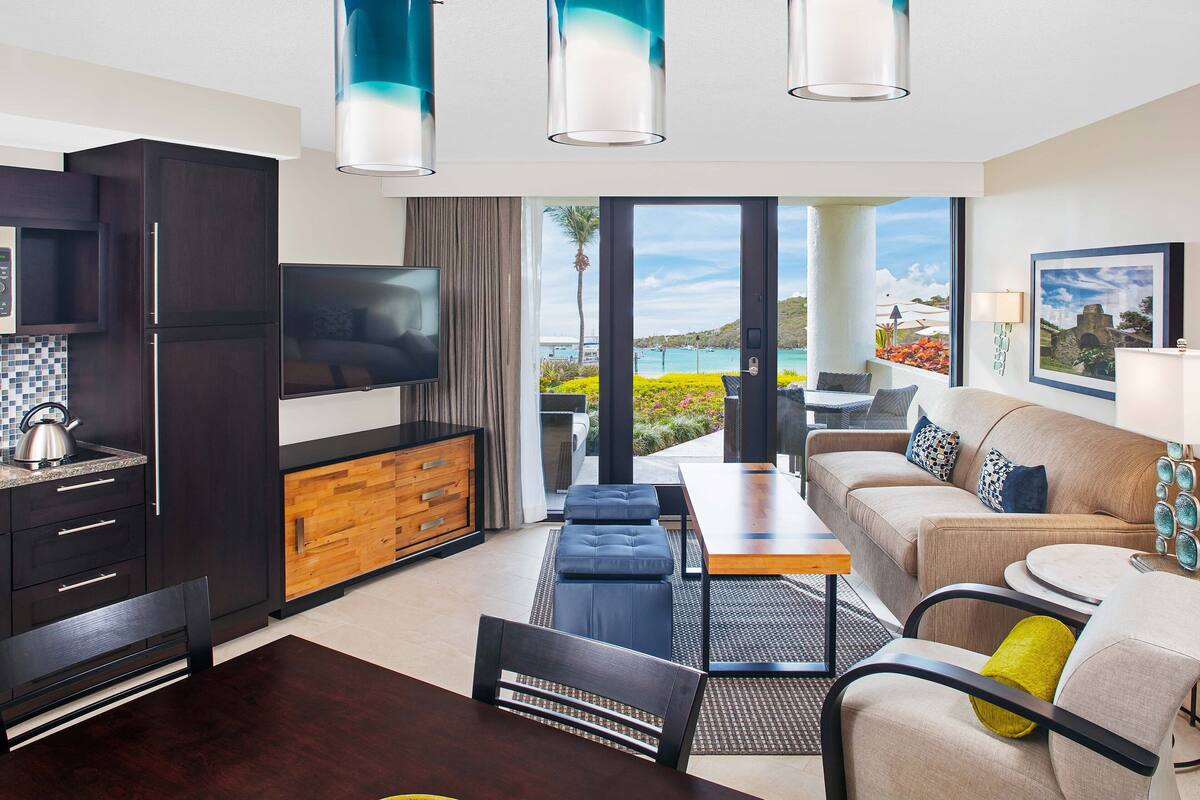 Una sala de estar moderna con sofá, mesa de centro y televisor. Cuenta con una zona de cocina con armarios y una mesa de comedor. La habitación tiene ventanas amplias con vistas al exterior.