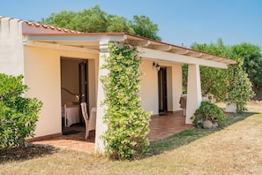 Un pequeño edificio de una sola planta con un porche cubierto, una puerta de acceso a una habitación y un pequeño jardín con un árbol y arbustos.
