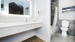 A modern bathroom with a white vanity, a large mirror, a handheld hair dryer, a white toilet, and a shower area. - Palace Inn Fairbanks (Houston)