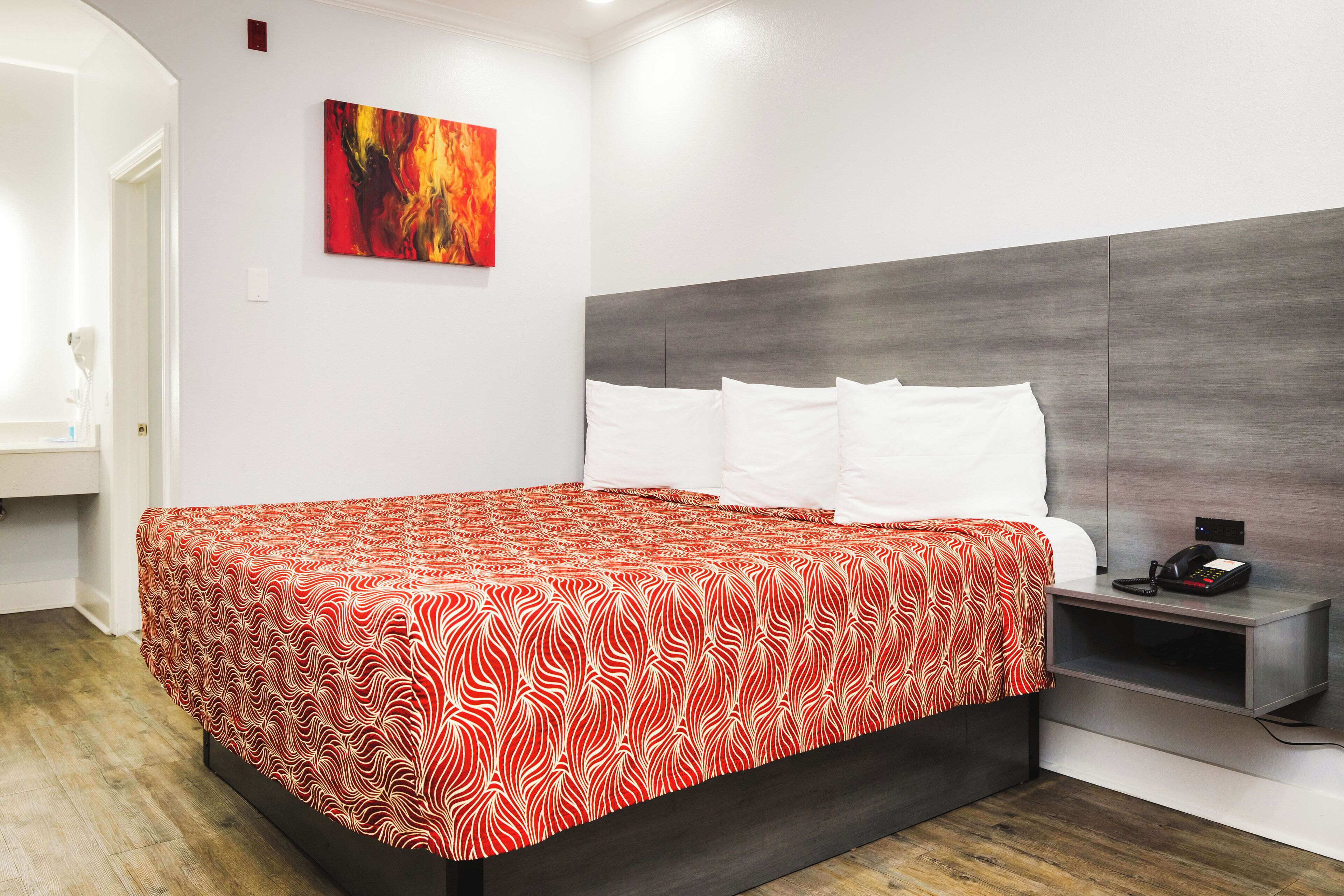 A modern hotel room with a large bed, white pillows, a red and white patterned bedspread, a grey headboard, a bedside table with a phone, and a painting on the wall.