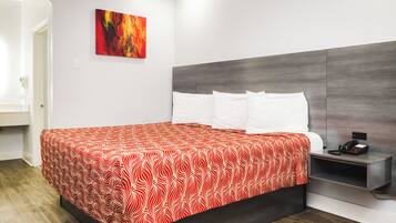 A modern hotel room with a large bed, white pillows, a red and white patterned bedspread, a grey headboard, a bedside table with a phone, and a painting on the wall.