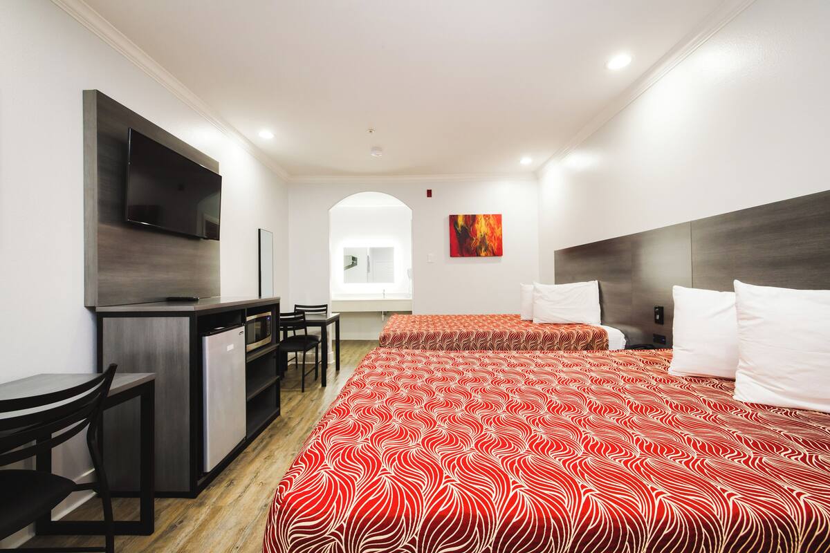 A hotel room with a kitchenette, two beds with red and white patterned bedding, a flat-screen TV mounted on the wall, and a painting on the wall.