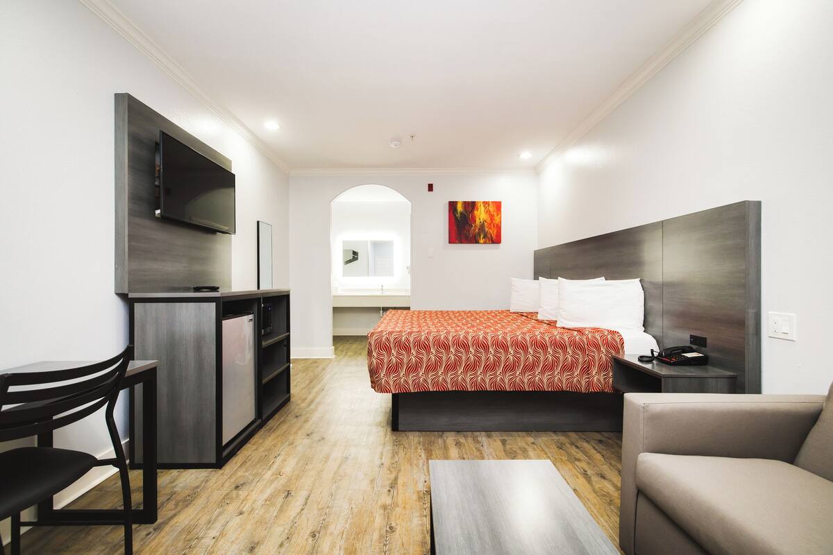 A compact hotel room with a bed, a desk, a chair, a TV mounted on the wall, and a painting on the wall.