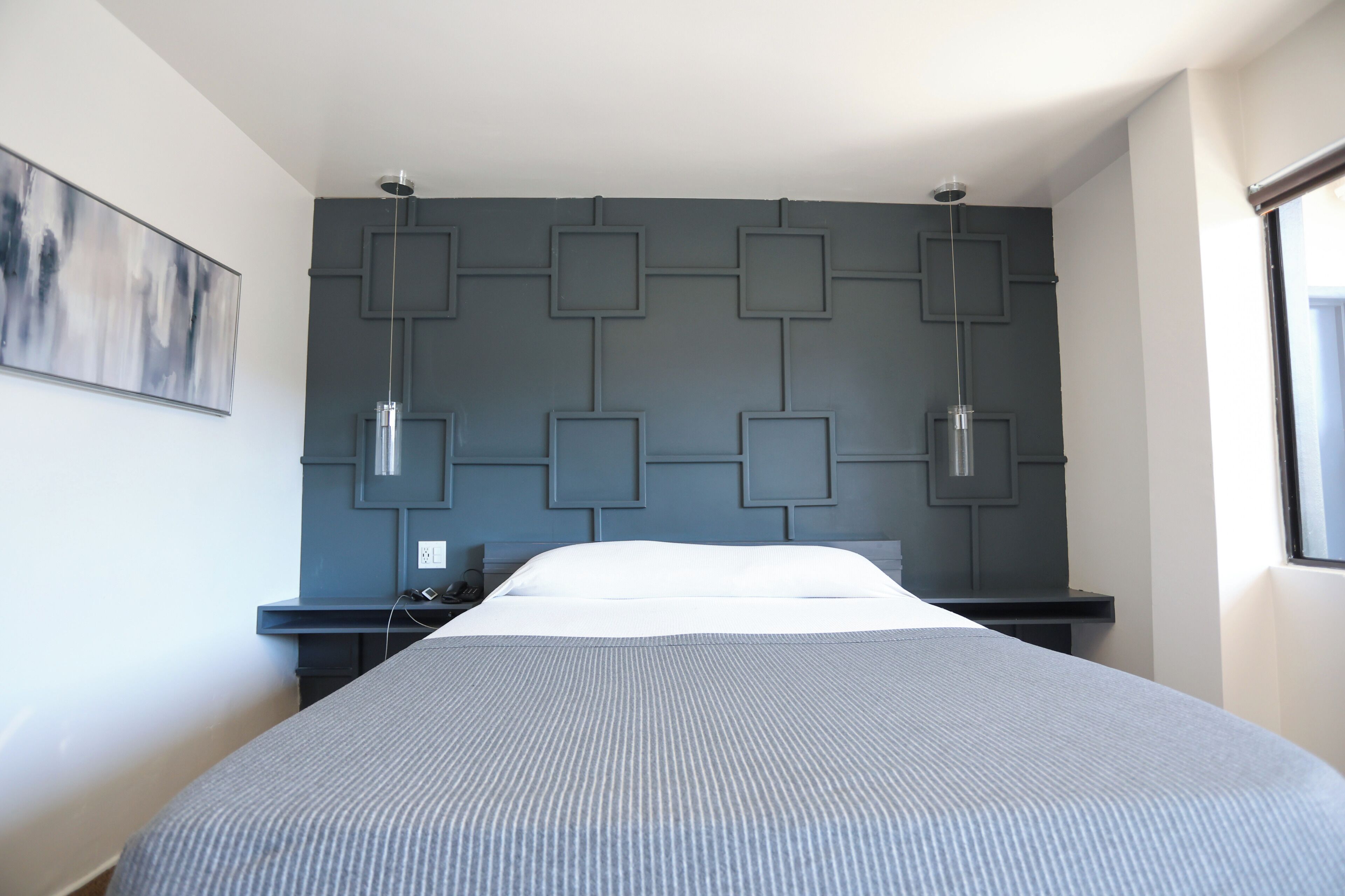 A modern hotel room with a large bed, a headboard with geometric patterns, a bedside table with a lamp, and a painting on the wall.