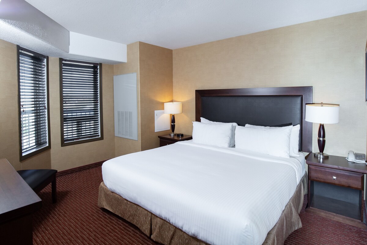 A hotel room with a large bed, two bedside tables with lamps, a desk, and a window with blinds.