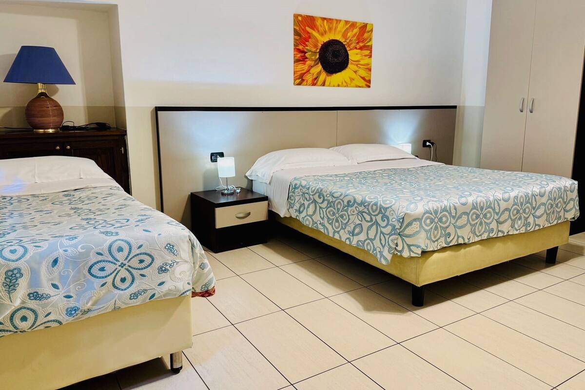 A hotel room with two beds, a nightstand with a lamp, a painting of a sunflower, and a wardrobe.