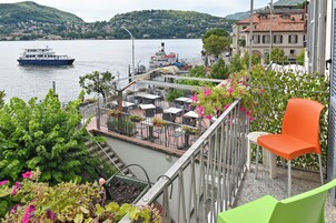 A terrace with outdoor seating overlooking a lake, a boat docked nearby, and a building with a red-tiled roof in the background.
