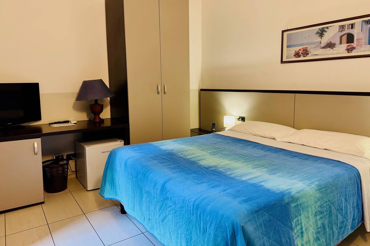 A hotel room with a bed, a desk with a lamp, a television, and a wardrobe.