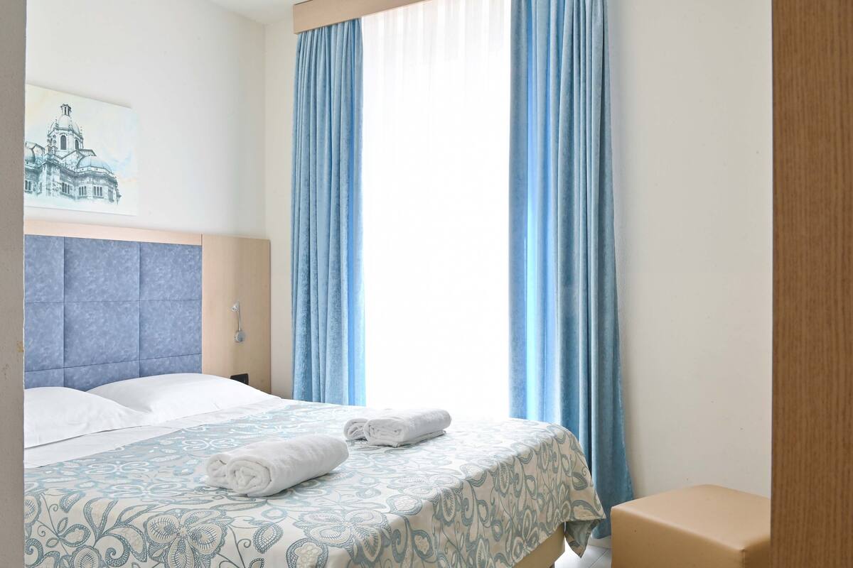 A hotel room with a bed, a headboard, a window with curtains, and a wall-mounted artwork.